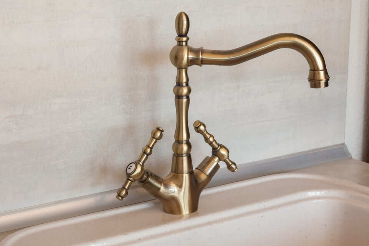 Brushed brass kitchen tap with cross handles and a curved spout above a ceramic sink.