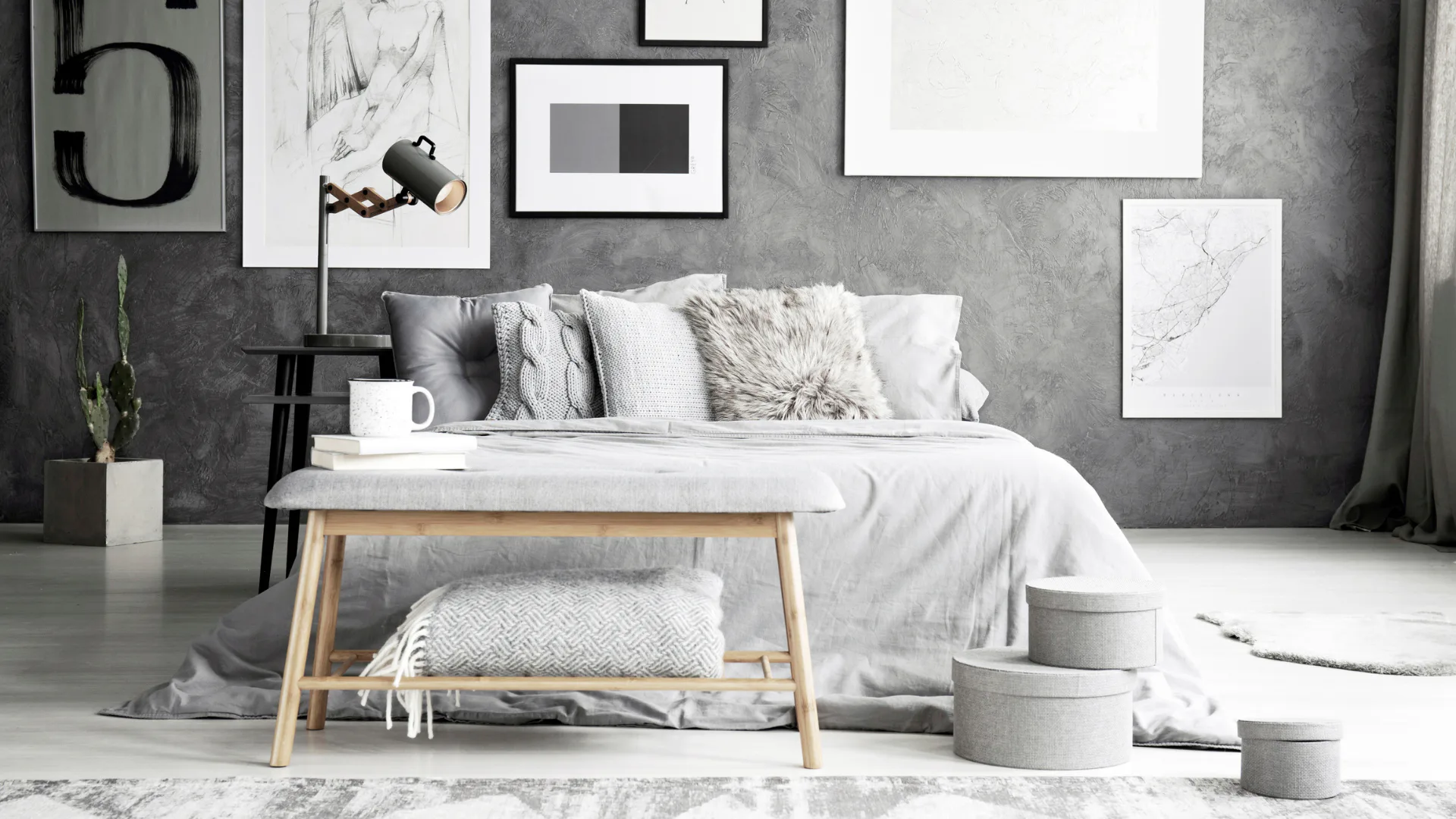 Minimalist bedroom featuring a white bed with gray and textured pillows, wooden bench with cushion, gray concrete accent wall, framed artwork, pendant lamp, potted plant, storage baskets, and neutr...