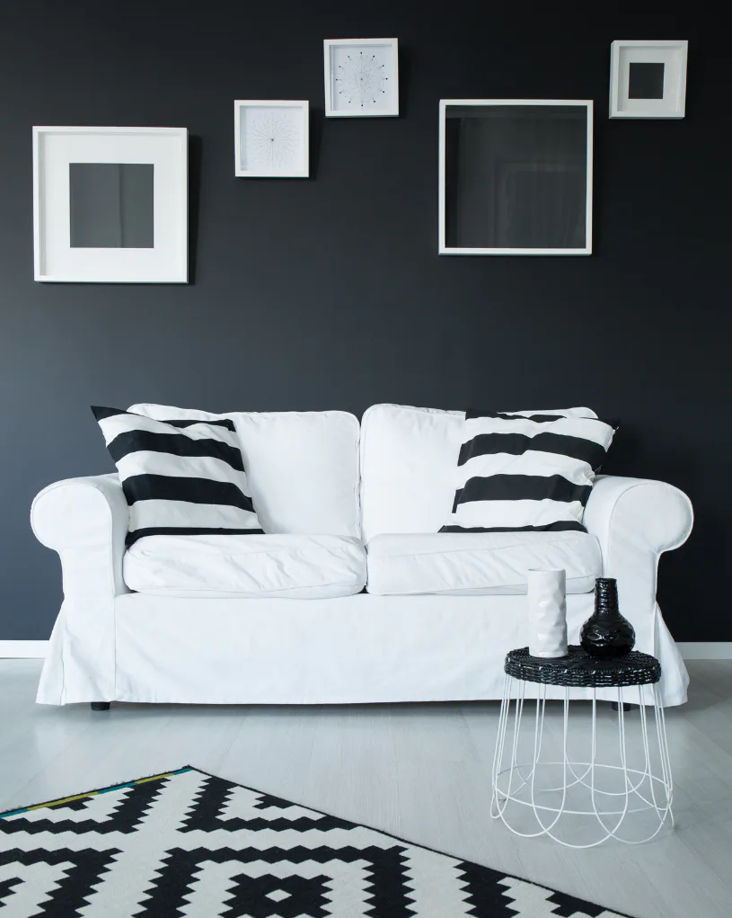 Minimalist living room featuring a white slipcovered sofa with black and white striped pillows against a charcoal wall, decorated with white-framed artwork and a geometric patterned rug