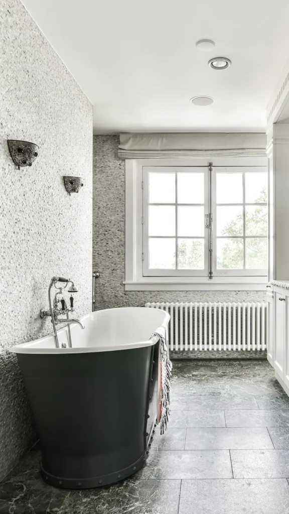 Elegant bathroom featuring a black freestanding soaking tub with white interior, white terrazzo tile walls, recessed ceiling lights, large window with grid panes overlooking trees, and white radiat...