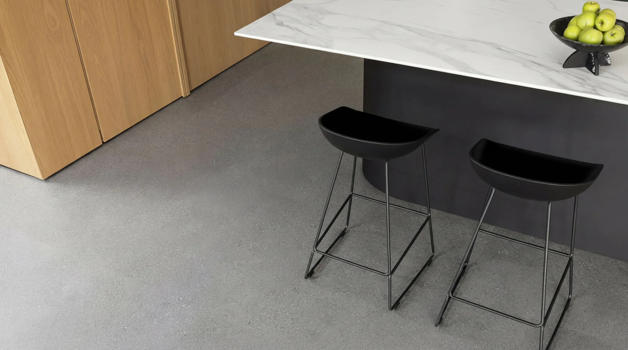 Contemporary kitchen featuring a marble-top island with black metal frame, two black bar stools with curved seats, wooden cabinetry, concrete flooring, and green apples in a decorative bowl.