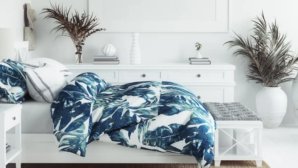 Bright coastal bedroom featuring a white dresser with blue and white botanical patterned bedding, dark palm fronds in glass vase, potted palm plant, stacked books, and minimalist wall décor.
