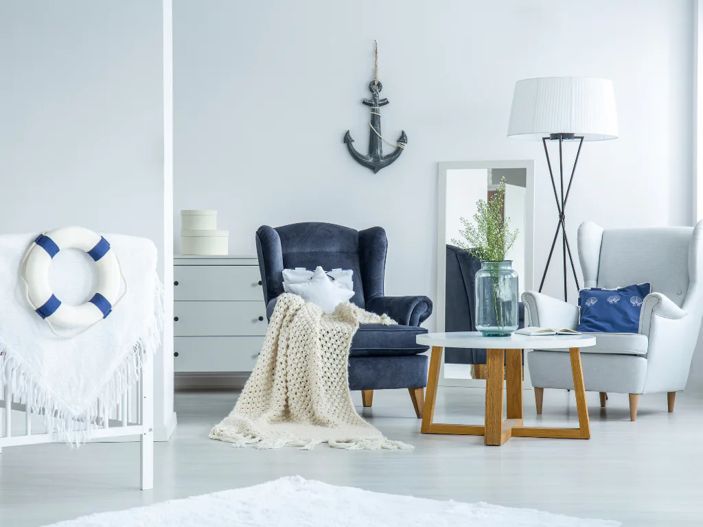 Bright coastal living room featuring a dark blue wingback chair with cream throw blanket, wooden coffee table, light gray armchair, white floor lamp, anchor wall decor, and blue-and-white life pres...
