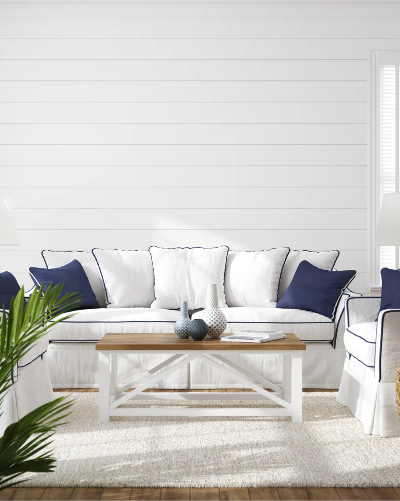 Bright coastal living room featuring a white upholstered sofa with navy and white pillows, white farmhouse coffee table with decorative vases, green potted plant, and shiplap accent wall with white...