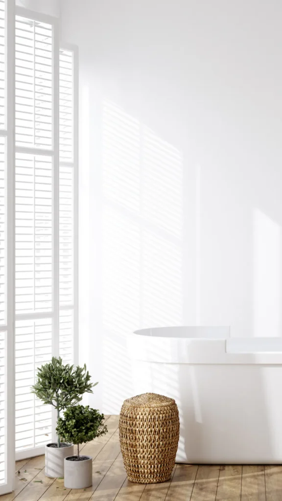 Bright minimalist bathroom interior featuring white plantation shutters, potted topiary plants in ceramic pots, a woven seagrass storage basket, and a freestanding white bathtub on wooden flooring