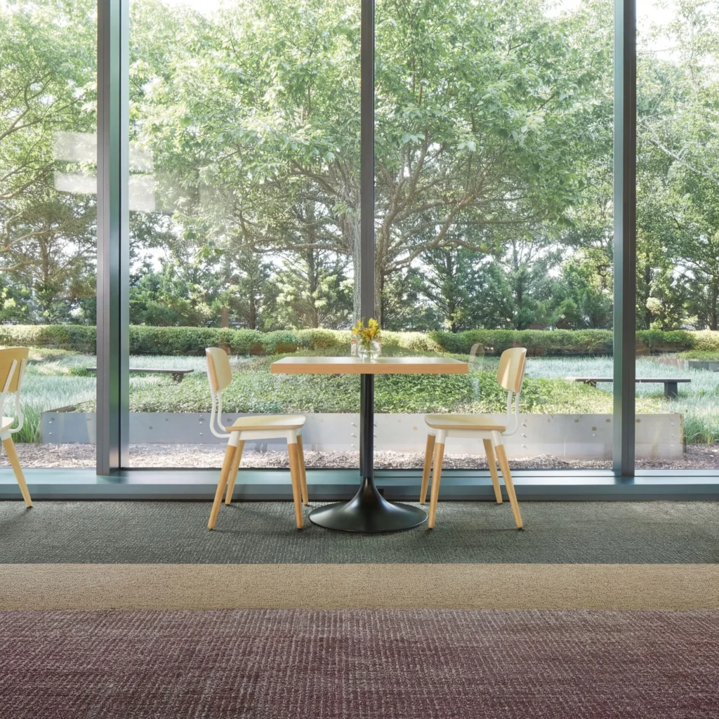 Modern office space with multi-colored commercial carpet flooring, wooden table with black pedestal base, light wood chairs, and floor-to-ceiling windows overlooking trees and landscaped grounds.