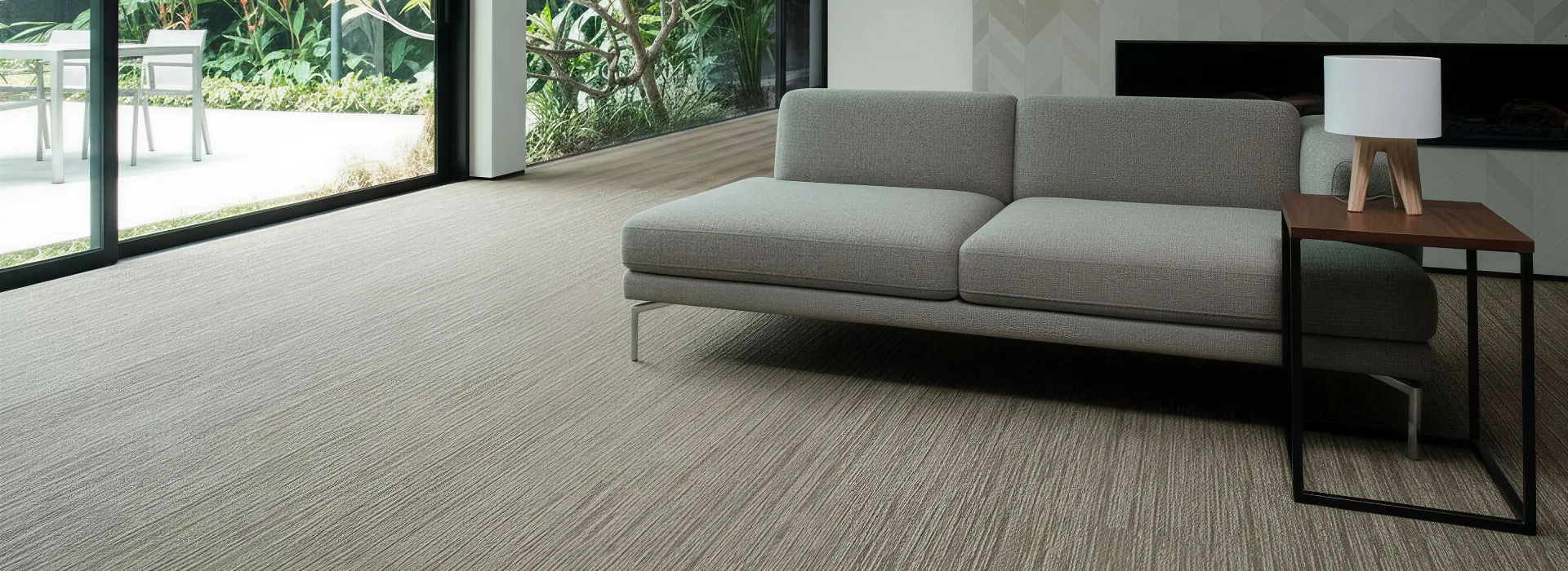 Contemporary gray upholstered sofa with metal frame and wooden side table on striped gray carpet tiles in minimalist office space with floor-to-ceiling windows overlooking garden