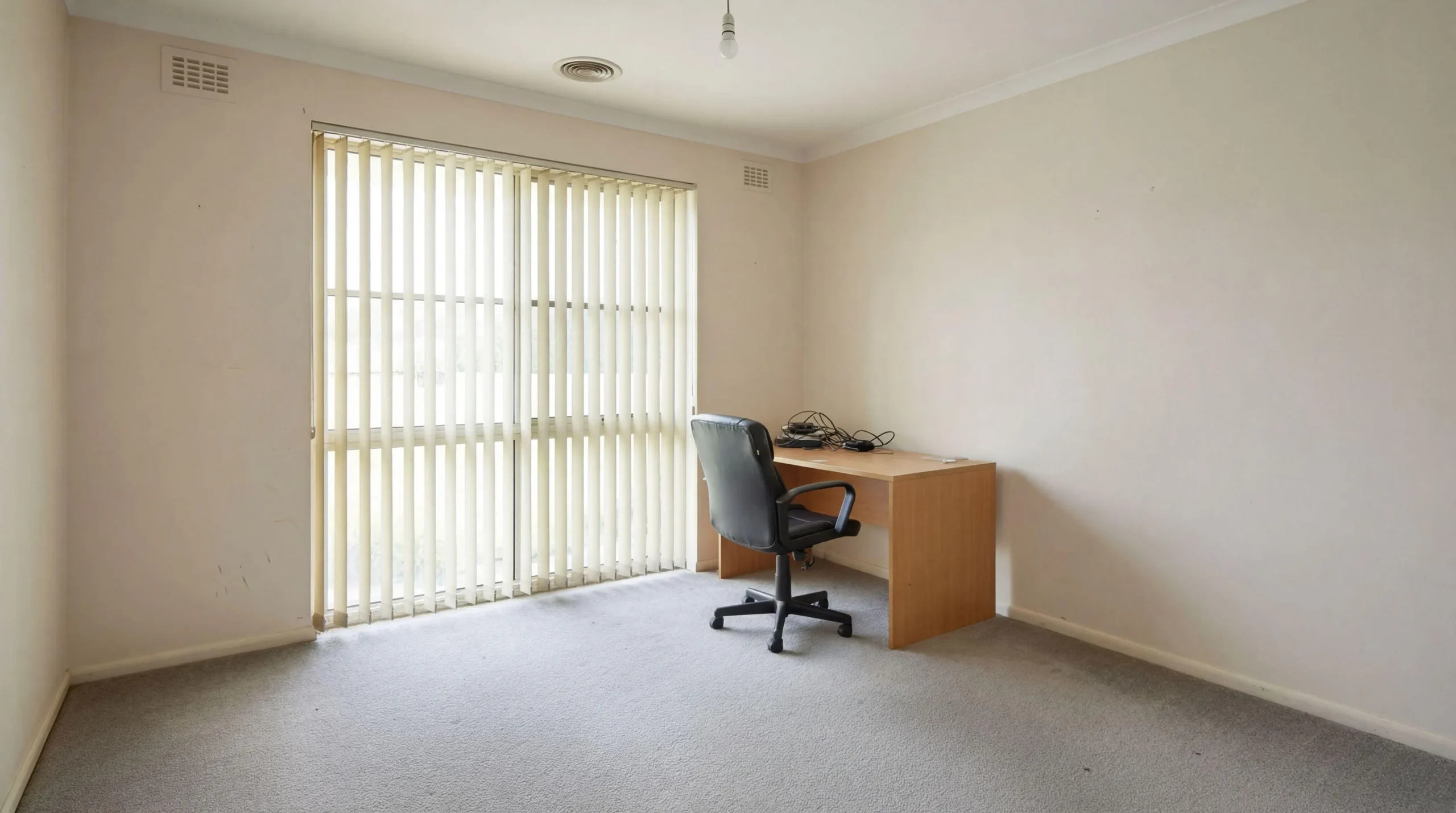 Empty office room with a light wood desk, black office chair, and cream vertical blinds covering a large window.