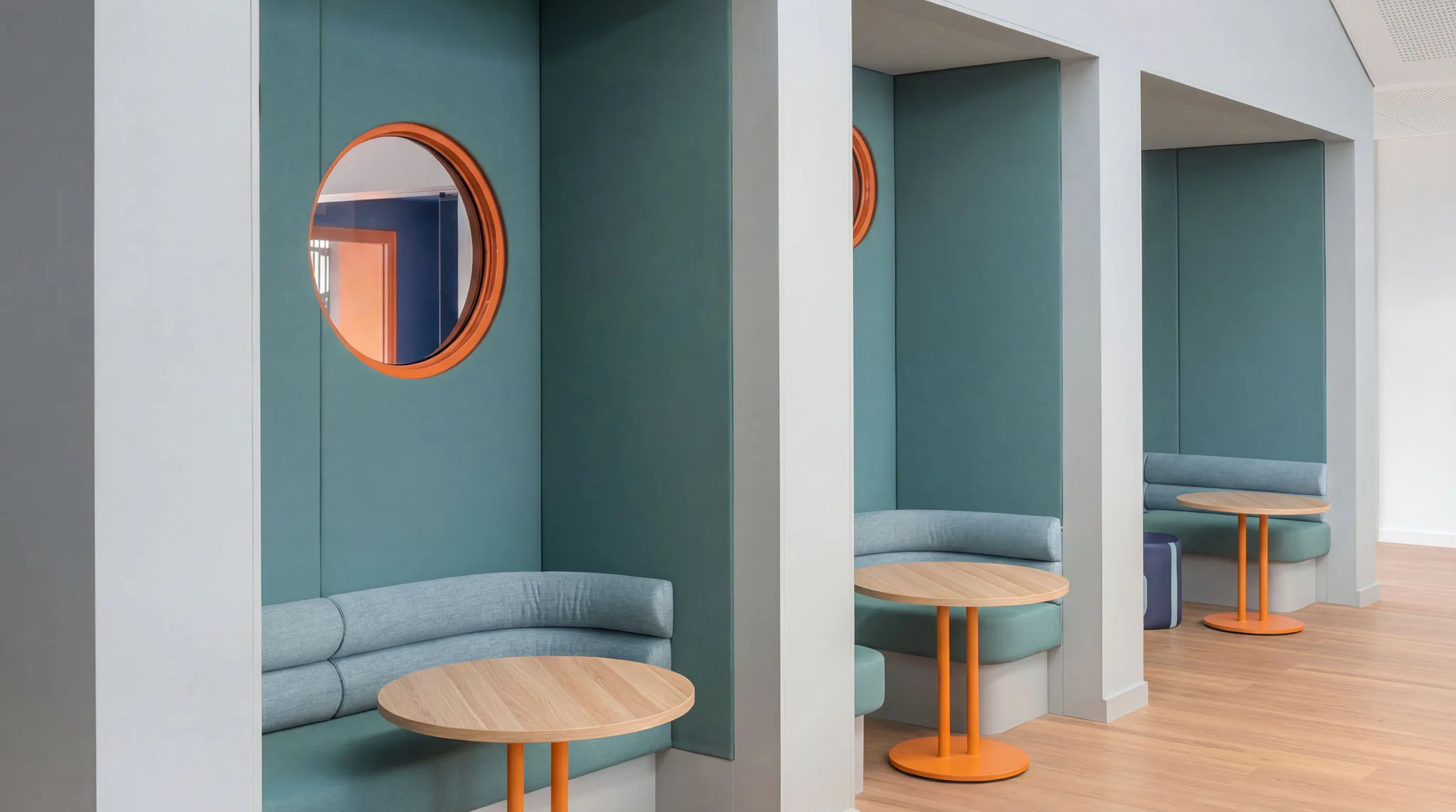 Contemporary office breakout area featuring teal curved alcove seating with gray upholstered bench seating, round wooden tables with orange metal bases, and circular orange-framed mirrors mounted o...