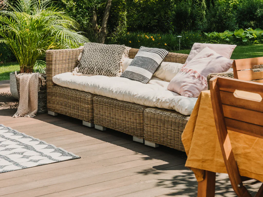 Natural wicker outdoor sofa with cream cushion and decorative pillows in geometric patterns, positioned on a wooden deck surrounded by lush green landscaping and tropical plants.