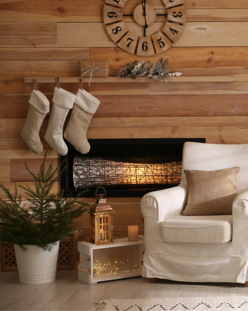 Cozy holiday living room featuring three cream stockings hung above a black fireplace with warm lighting, wooden wall clock, evergreen garland, white armchair with tan pillow, potted greenery, and ...