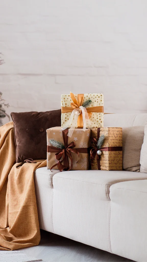 Three wrapped presents stacked on a cream-colored sofa, featuring gold and brown kraft paper with ribbon bows and evergreen sprigs, alongside brown and gold throw pillows and a mustard blanket