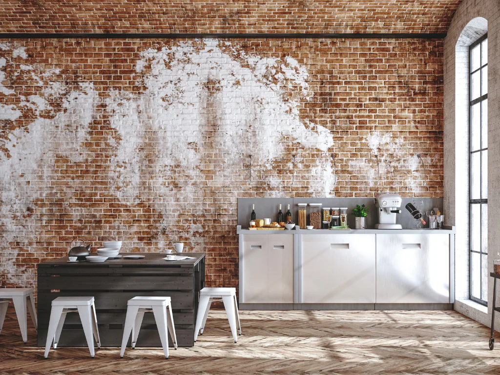 Industrial modern kitchen featuring distressed red brick walls, white metal bar stools, dark wood island counter, stainless steel cabinets with gray backsplash, and tall windows providing natural l...
