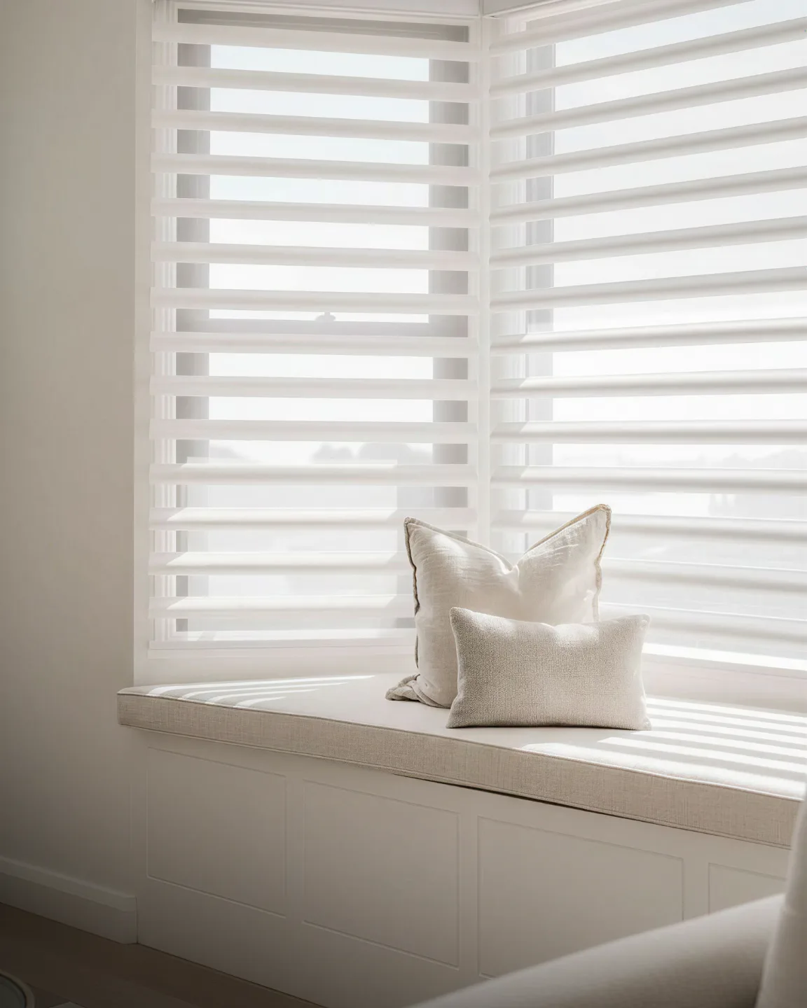 Bright white motorized shutters frame a minimalist window seat with cream-colored cushion and two linen pillows, bathed in natural light filtering through partially open horizontal slats.
