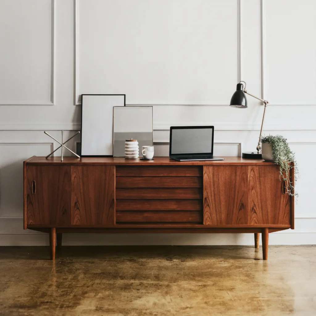 Walnut wood mid-century credenza with tapered legs supporting a workspace featuring a laptop, framed mirrors, desk lamp, potted plant, and decorative accessories against a white paneled wall.