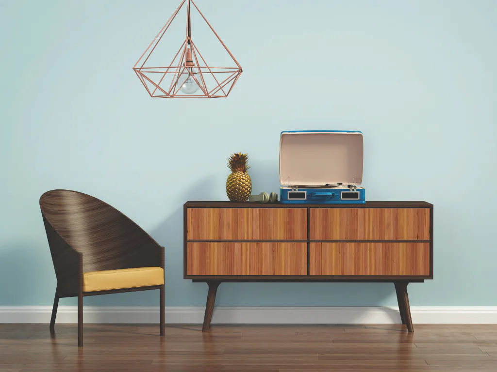 A mid-century modern living room featuring a wooden dresser with four drawers, a blue record player on top, a curved chair with yellow cushion, a hanging geometric light fixture, and a pineapple de...