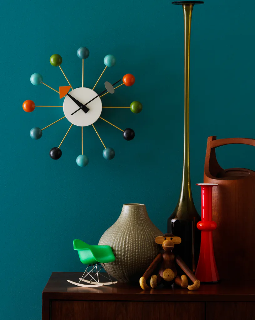 Styled mid-century modern shelf display featuring a sunburst atomic clock with colored spheres, textured ceramic vase, green Eames-style chair, brass floor lamp, wooden articulated figurine, and re...