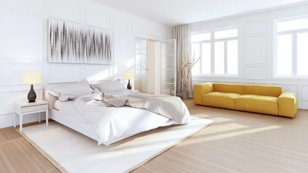 Bright modern bedroom featuring a white bed with gray bedding, white nightstand with lamp, abstract wall art, and a bold yellow modular sofa positioned near large windows with natural light floodin...