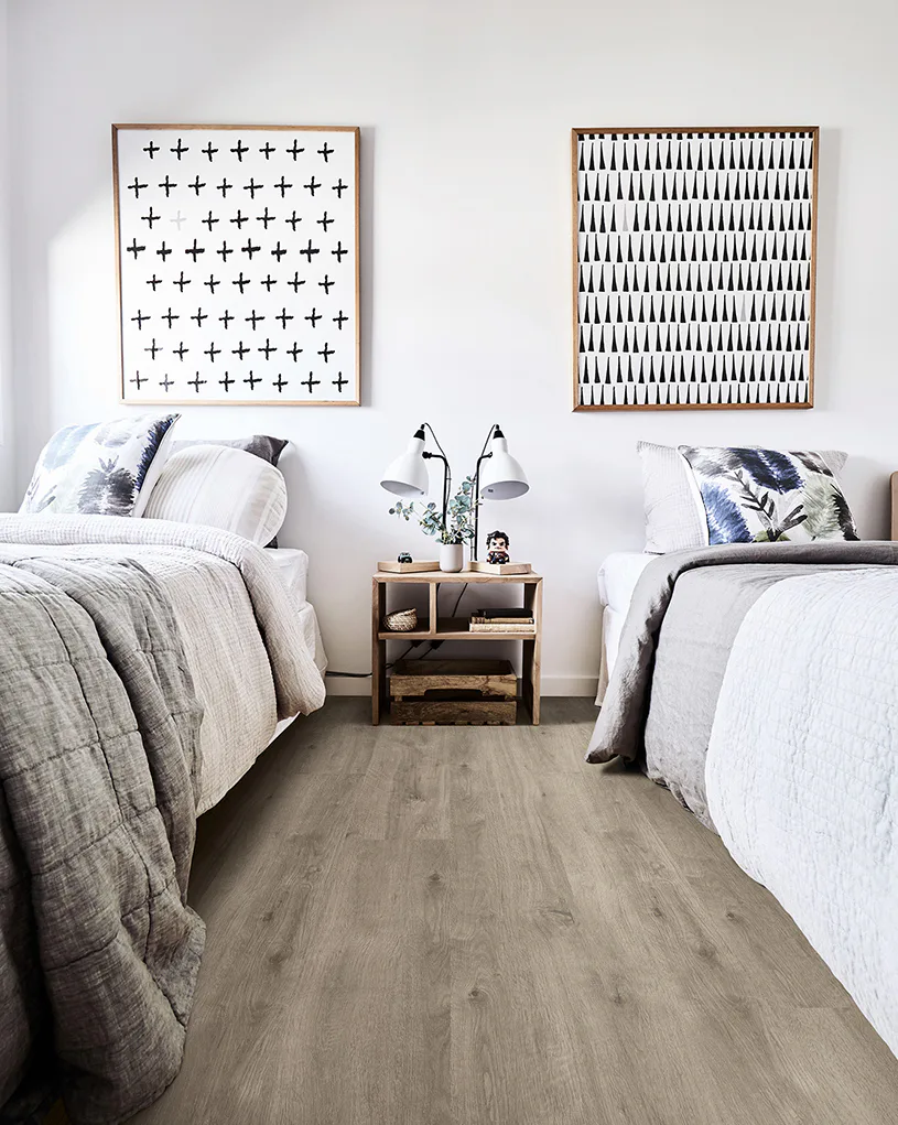 Modern bedroom featuring two twin beds with white linens and blue accent pillows, separated by wooden nightstand. Gray laminate flooring spans the room with geometric wall art above each bed.