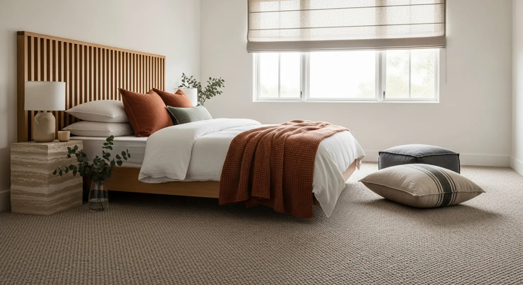 Bright modern bedroom featuring beige wool carpet, wooden slatted headboard, white bedding with rust-colored throw and pillows, stone nightstand with plants, and natural window light