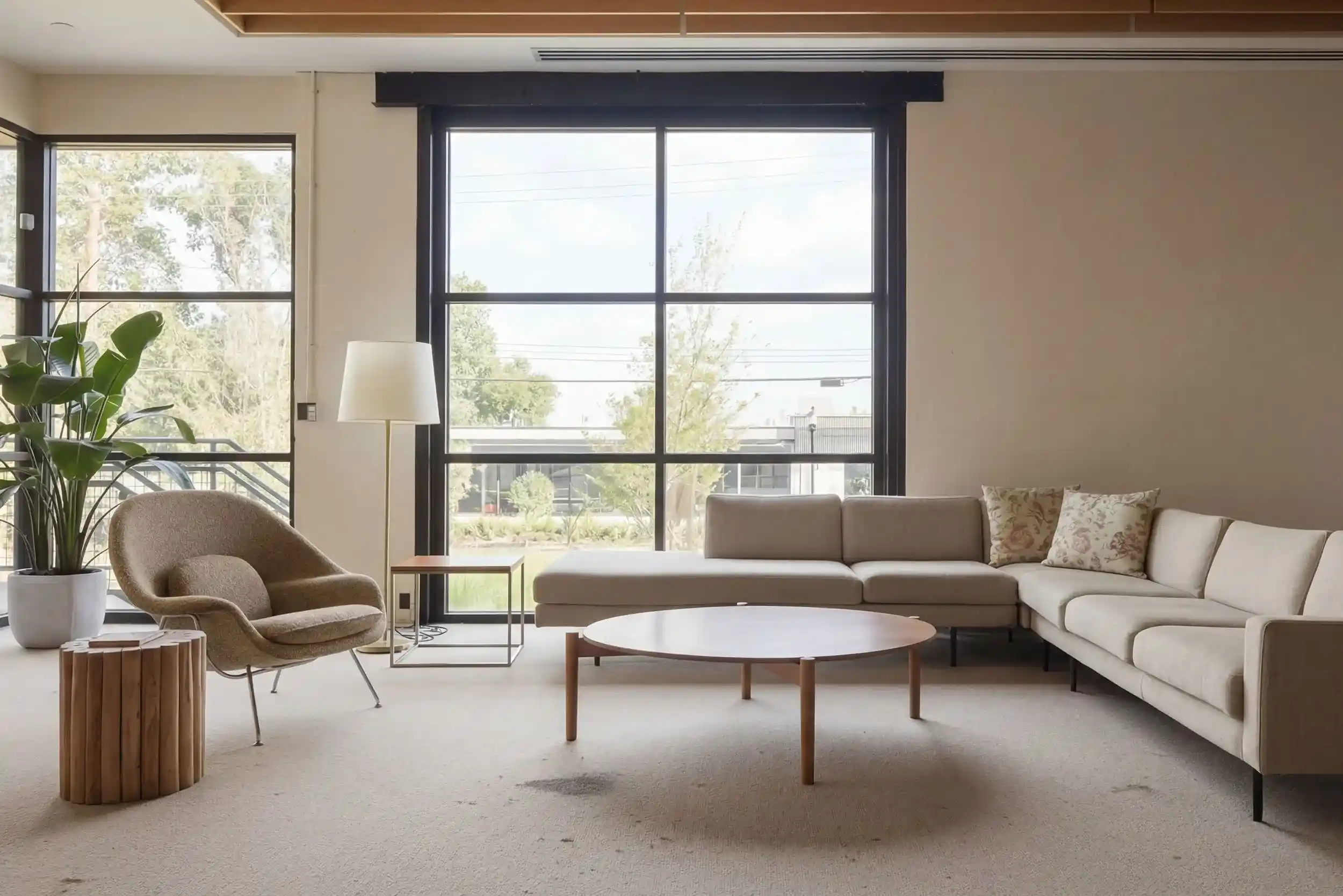 A modern living room featuring a large, light beige sectional sofa and a round wooden coffee table. Natural light streams through the large window.