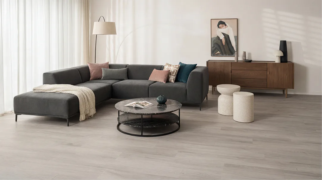 Contemporary living room featuring a charcoal gray sectional sofa with colorful accent pillows, a round black metal coffee table, cream pouf seating, wooden sideboard, arc floor lamp, and light woo...
