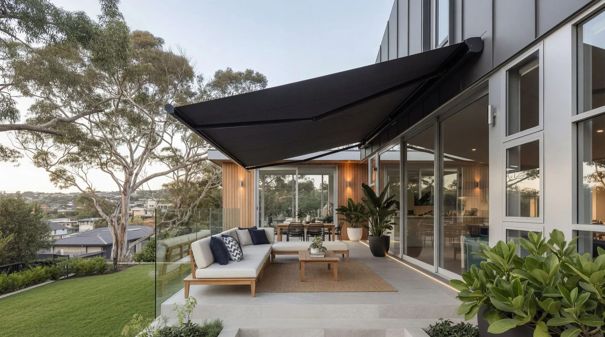 Large black triangular awning providing shade over contemporary outdoor patio featuring light wood furniture with gray cushions, glass railings, and lush green plants