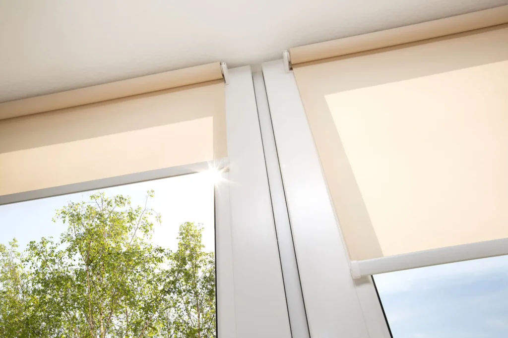 Two cream-colored roller blinds mounted on a wooden track inside a bright room with large windows overlooking green foliage and blue sky, demonstrating contemporary window covering installation.