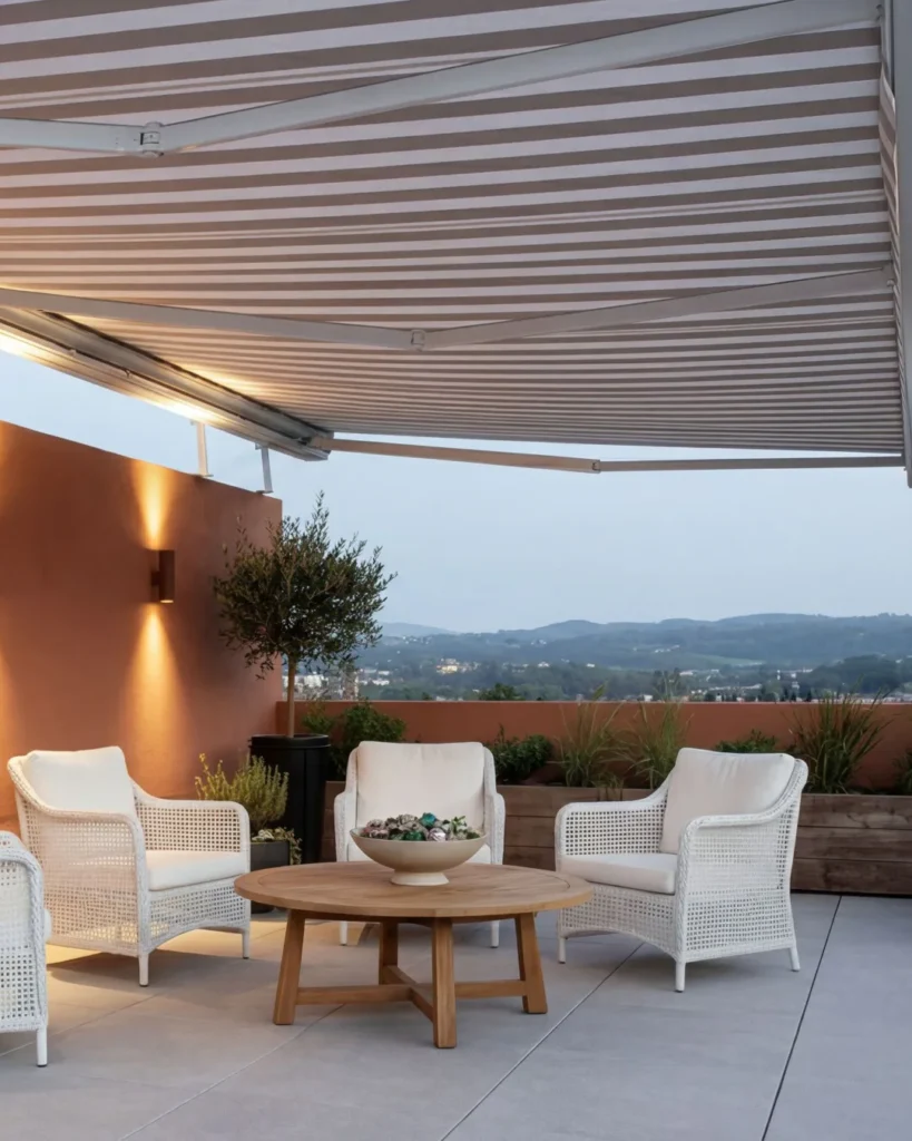 Beige and brown striped retractable awning extended over modern patio seating area with white wicker chairs around wooden coffee table, overlooking mountain landscape