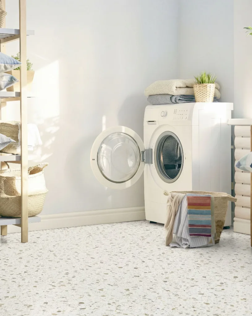 Bright laundry room with white sheet vinyl flooring featuring speckled texture, white front-load washing machine, stacked folded towels, woven storage baskets, and potted plant on shelving