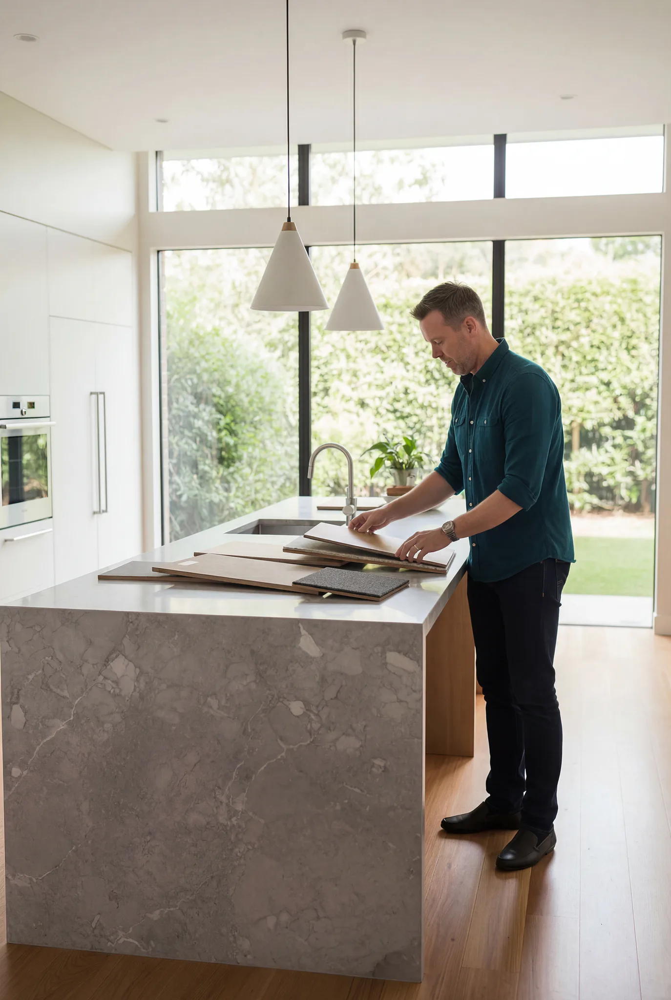 Man in navy shirt examining flooring samples at modern kitchen island with pendant lights and garden views
