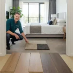 Smiling flooring professional in teal shirt crouching beside wood flooring samples in bedroom, pointing to carpet and rug display with modern bed and natural light background