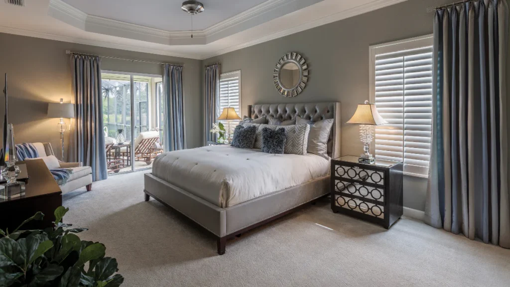 Elegant traditional bedroom featuring a gray upholstered bed with tufted headboard, white bedding, decorative pillows, two nightstands with table lamps, ceiling fan, and shuttered windows with blue...