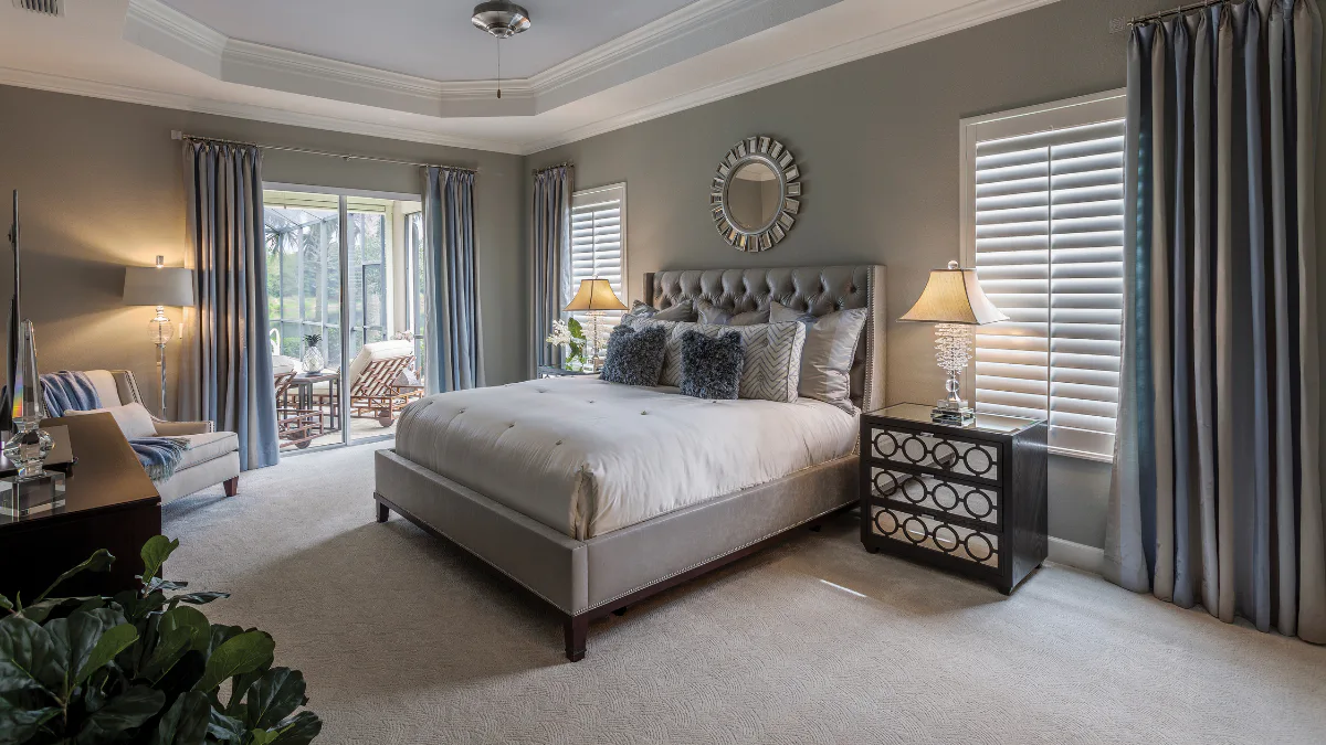 Elegant traditional bedroom featuring a gray upholstered bed with tufted headboard, white bedding, decorative pillows, two nightstands with table lamps, ceiling fan, and shuttered windows with blue...