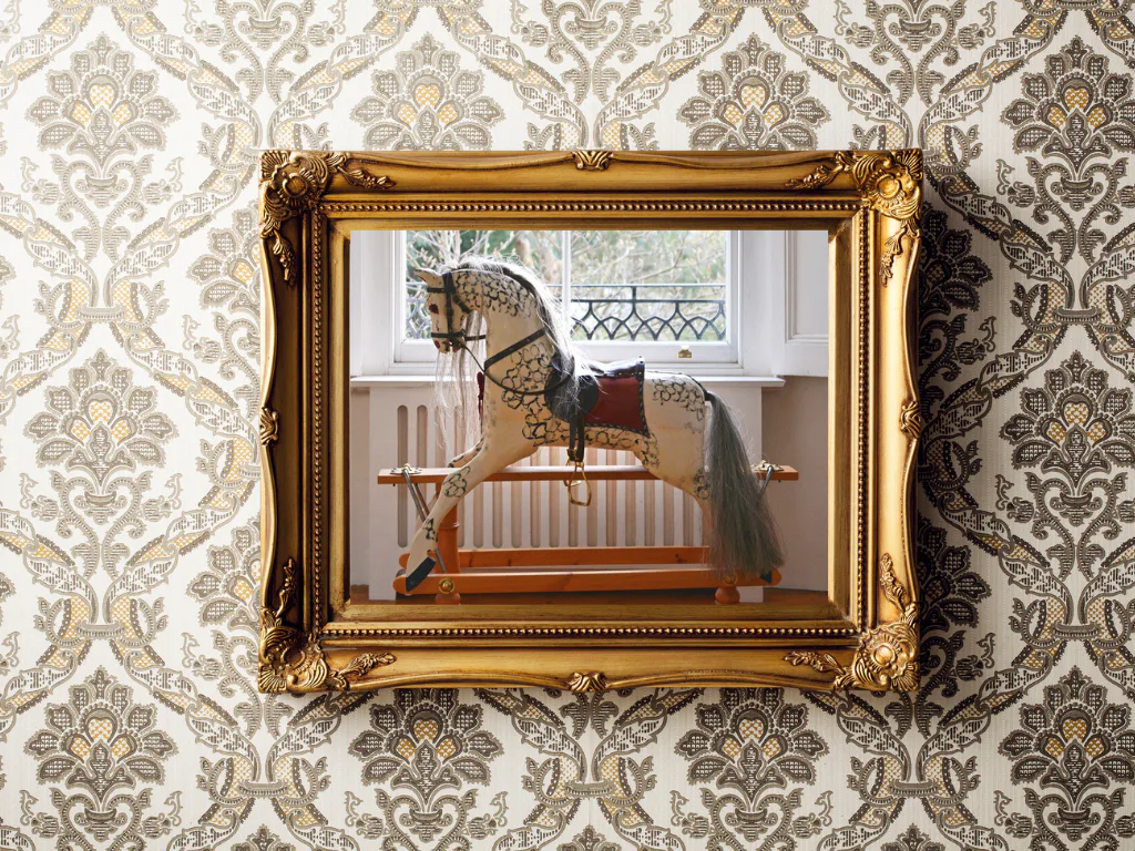 A decorative wooden rocking horse with ornate gold and black saddle detailing stands against a cream radiator beneath a sunlit window, all framed by an elaborate gold-leaf picture frame mounted on ...