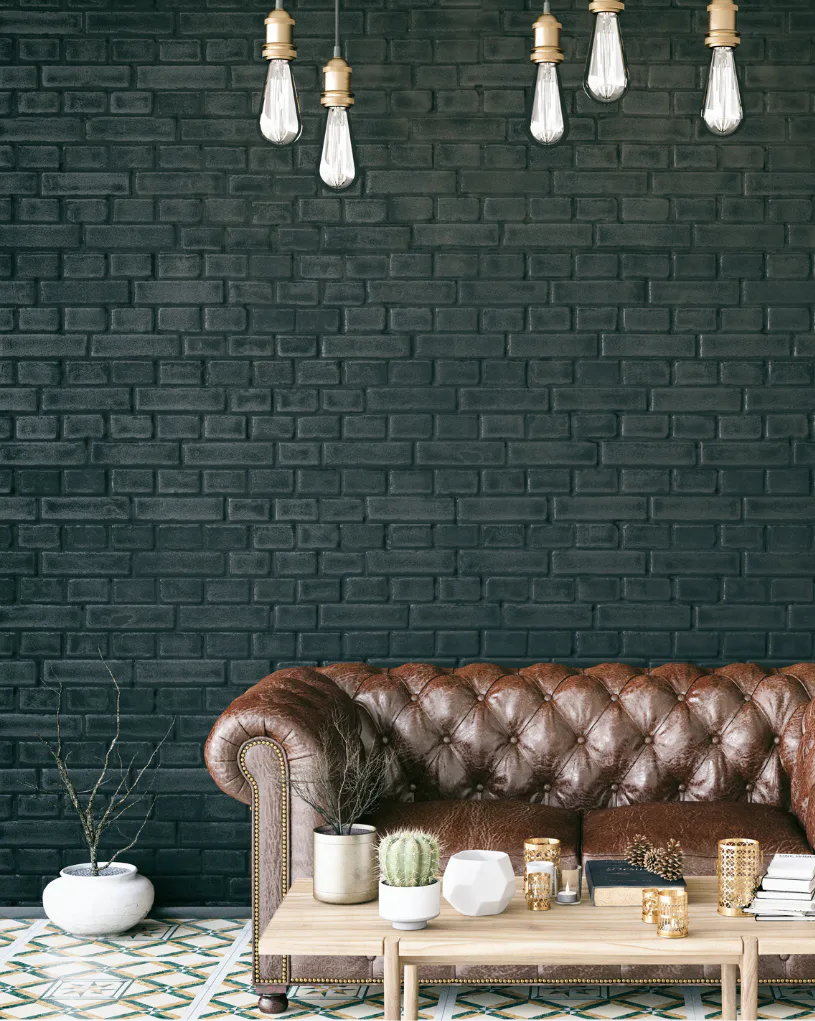 Brown leather Chesterfield sofa against dark green painted brick wall with hanging Edison bulb pendant lights, wooden table with potted cactus and decorative items