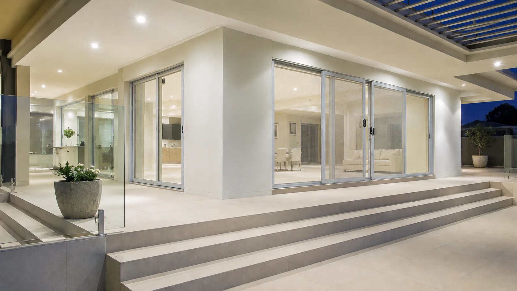 Illuminated modern home exterior featuring multiple aluminum-framed sliding glass doors with integrated security screens, concrete stepped patio, recessed lighting, and planted pot