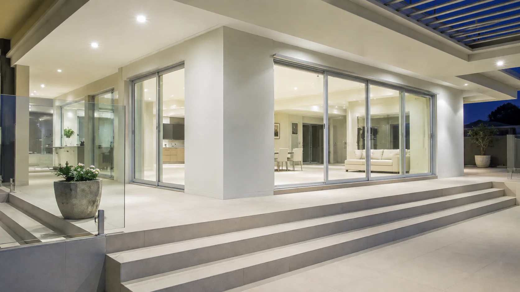 Contemporary home exterior featuring large frameless glass sliding doors opening onto a spacious patio with stepped entry, white columns, recessed lighting, and interior living space visible throug...